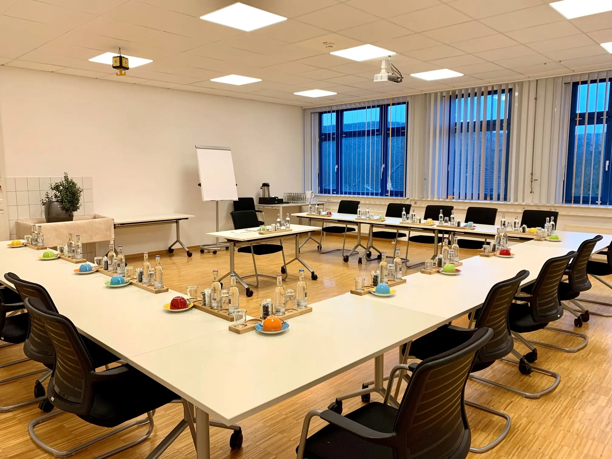 Gestaltungsraum. Tagungsraum. Tische in U-Form angeordnet. Die Tische sind gedeckt mit jeweils 2 Wasserflaschen und 2 Gläsern, sowie einer Kaffeetasse. Im U steht ein Tisch für einen Redner mit gleicher Bestückung. Hinter dem redner Tisch befindet sich ein Flipchart.