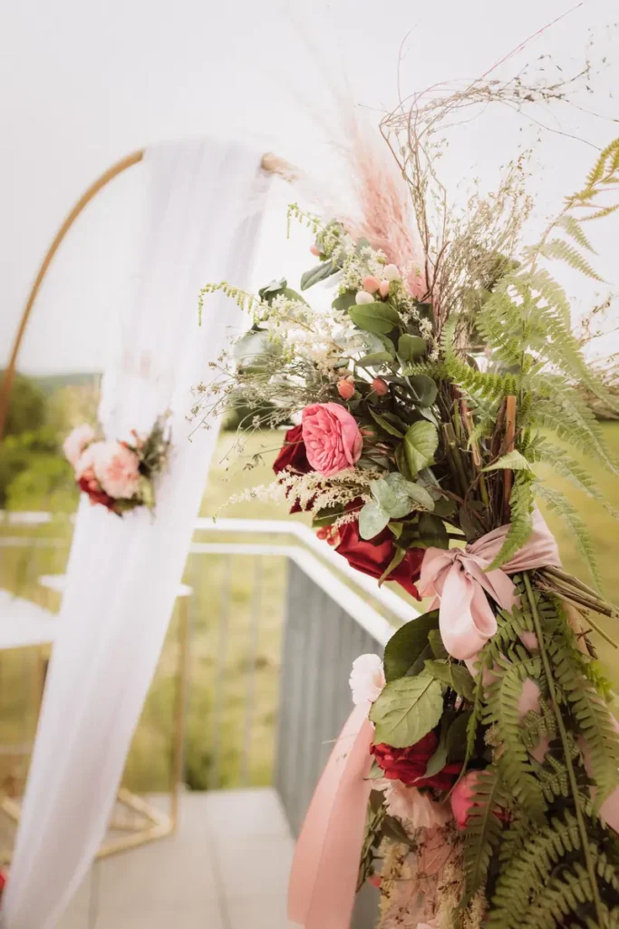Die Nahaufnahme zeigt eine stilvolle Hochzeitsdekoration mit einer Blumenarrangement, das frische rosa und rote Blumen umfasst. Das Arrangement ist reichhaltig und wird von grünen Blättern und zarten Gräsern umrahmt. Eine weiße, transparente Stoffbahn hängt elegant im Hintergrund, während eine geschwungene goldene Struktur als Rahmen dient. Im Hintergrund sind sanfte, grüne Wiesen zu sehen, die eine romantische Atmosphäre schaffen. Die Verwendung von B
