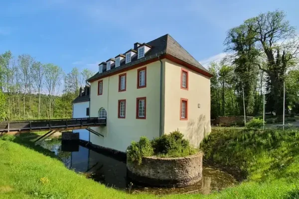 Eine malerische Ansicht der Wasserburg Hainchen. Schräge Ansicht mit dem Wassergraben und der Brücke zum Eingang., das sich am Ufer eines ruhigen Gewässers befindet. Das Haus hat eine hellgelbe Außenfarbe mit roten Fensterrahmen und einem Satteldach. Vor dem Gebäude führt eine Holzbrücke über den Wasserlauf. Im Vordergrund sind grüne Wiesen und kleine Sträucher zu sehen, die dem Bild eine natürliche Atmosphäre verleihen. Im Hintergrund wachsen hohe Bäume, die die Umgebung einrahmen.