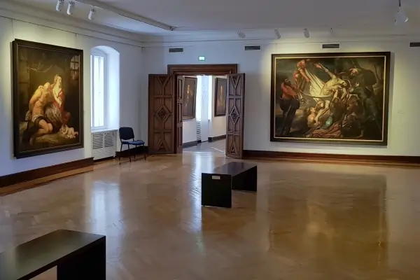 Das Bild zeigt einen hellen Ausstellungsraum des Siegerlandmuseums im Oberen Schloss mit Holzboden. An den Wänden hängen zwei große Gemälde, eines links, das eine Darstellung von Personen in emotionaler Interaktion zeigt, und ein weiteres rechts mit biblischem Motiv und dramatischer Komposition. In der Mitte des Raums steht ein minimalistischer, schwarzer Tisch. Auf der linken Seite ist ein schwarzer Stuhl sichtbar, während in der Hintergrunderöffnung eine weitere Galerie sichtbar ist.