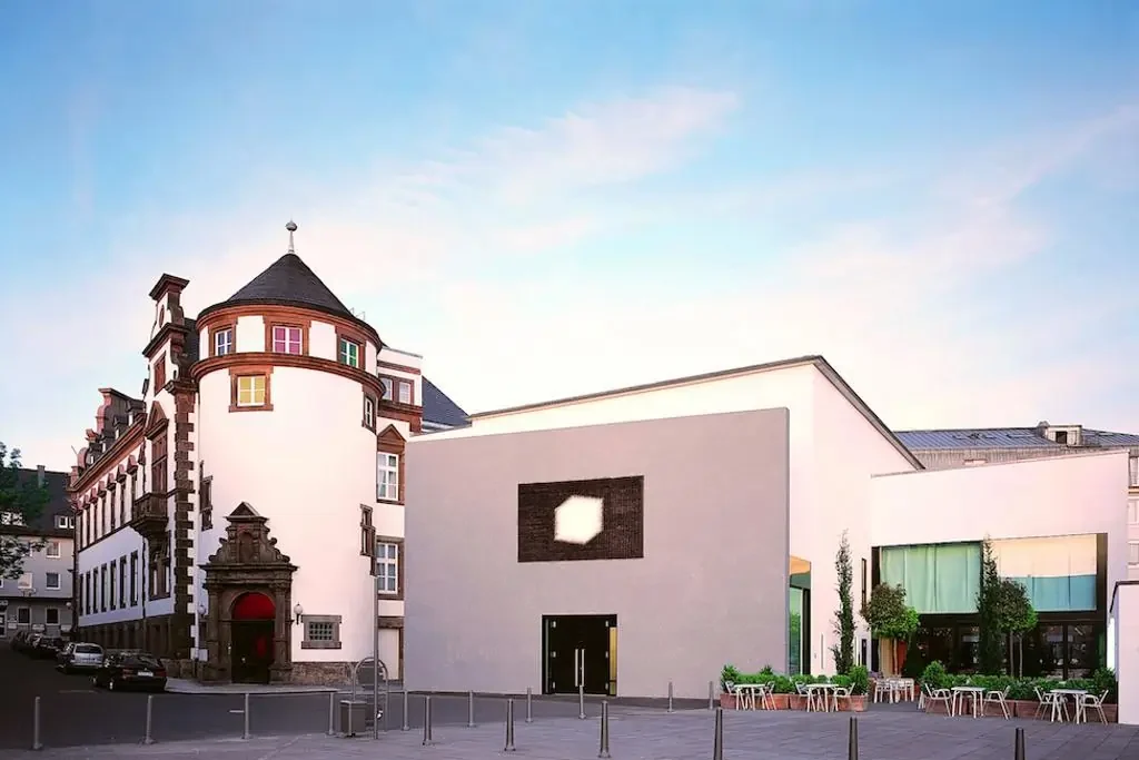 Außenansicht des Museums für Gegenwartskunst. Das Bild zeigt eine moderne Architektur im Kontrast zu einem historischen Gebäude. Links im Bild steht ein altes, mehrstöckiges Gebäude mit einem runden Turm und bunten Fenstern. Die Fassade ist aus traditionellem Stein und Holz gestaltet. Rechts daneben befindet sich ein zeitgenössisches, minimalistisches Gebäude mit einer glatten grauen Außenwand und großen Fensterfronten. Im Vordergrund sind Tische und Stühle auf einem Platz angeordnet, umgeben von Pflanzkästen