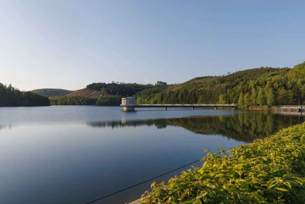 Ein Panorama der Obernautalsperre. Der klare See spiegelt die umliegenden grünen Hügel und den blauen Himmel wider. Am rechten Bildrand befindet sich ein gepflegtes Ufer mit üppigem, grünem Bewuchs. In der Bildmitte erhebt sich ein kleiner Pavillon oder Aussichtsturm auf einer langen Brücke, die über das Wasser führt. Im Hintergrund sind bewaldete Berge zu sehen, deren sanfte Konturen die friedliche Landschaft vervollständigen.