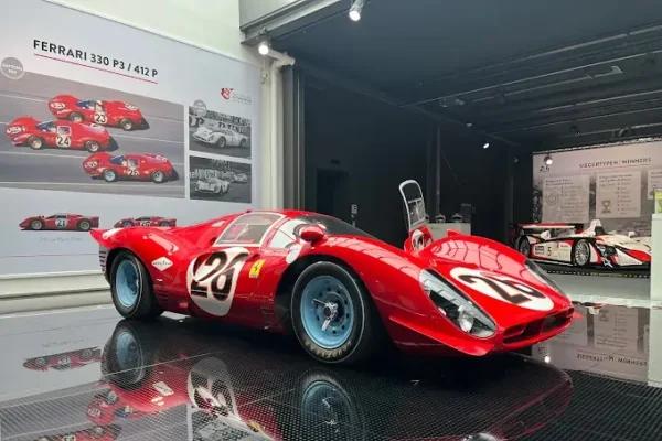 Ein roter Ferrari 330 P3 steht im Fokus des Bildes, mit der Nummer 26 auf den Türen. Der Sportwagen hat eine aerodynamische Form und glitzernde, silberne Felgen. Im Hintergrund ist eine Wand mit historischen Schwarz-Weiß-Fotos von Rennwagen zu sehen. Die Ausleuchtung des Raumes hebt die Details des Fahrzeugs hervor und schafft eine ansprechende Präsentation. Der Boden ist glänzend und reflektiert das Licht.