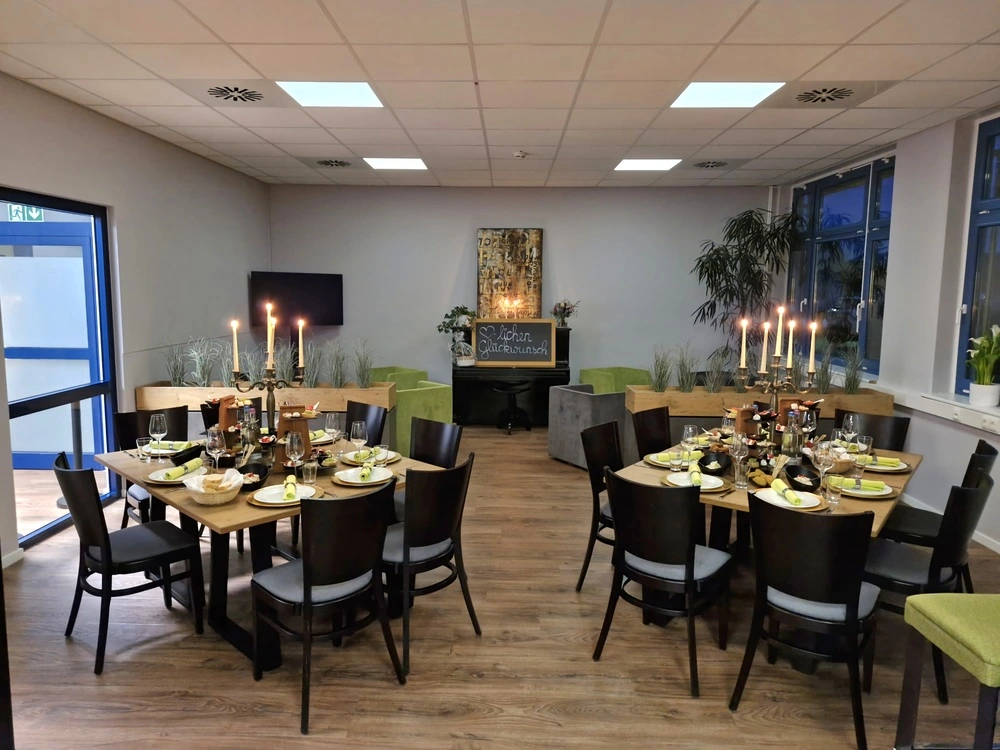 Das Bild zeigt einen ansprechend dekorierten Raum, der für eine festliche Veranstaltung oder ein Dinner vorbereitet ist. Zwei runde Esstische sind liebevoll eingedeckt mit Geschirr, Gläsern und Servietten. Auf den Tischen befinden sich auch verschiedene Speisen, darunter wohl appetitliche Snacks. Um die Tische stehen Stühle mit dunklem Holz und einer gepolsterten Sitzfläche. In der Mitte der Tische stehen Kerzenleuchter mit brennenden