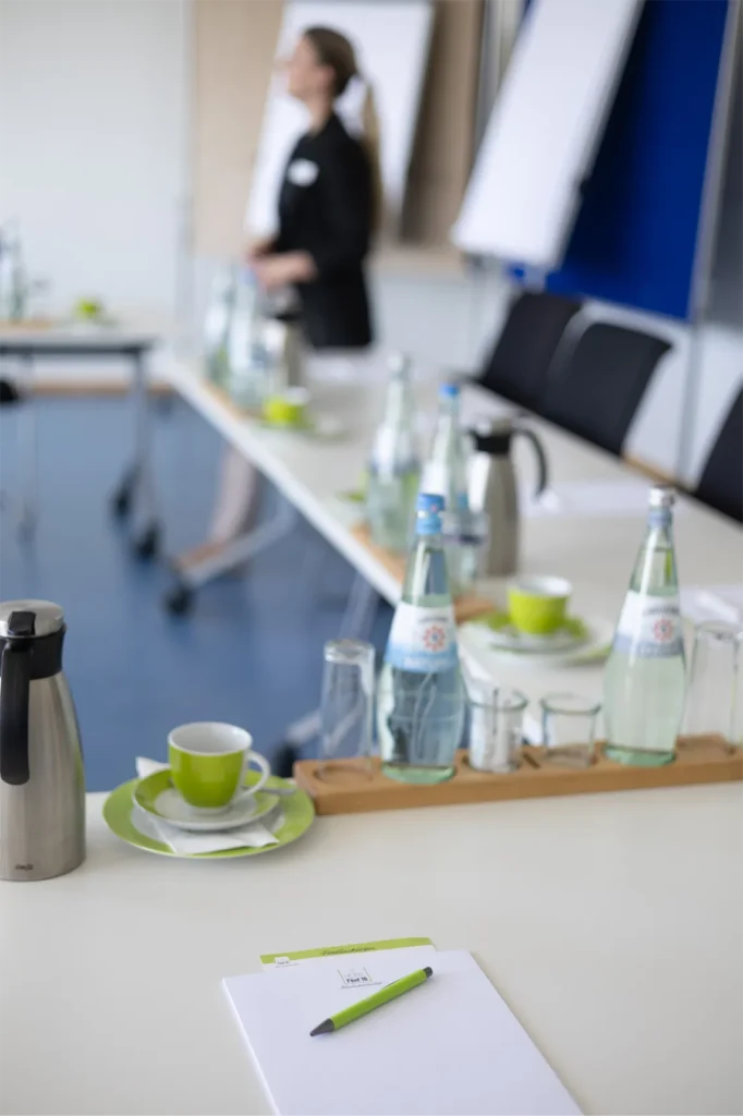 Ein professionell gedeckter Konferenzraum im HotelFünf 10 mit einem langen Tisch, auf dem verschiedene Getränke und Snacks bereitstehen. Im Vordergrund liegt ein Notizblock mit einem grünen Stift. Die Tischdekoration umfasst Wassergläser, Mineralwasserflaschen und kleine grüne Kaffeetassen auf passenden Untertellern. Im Hintergrund ist eine Frau teilweise unscharf zu sehen, die einen Vortrag hält, während Flipcharts und ein blauer Wandbereich sichtbar sind.