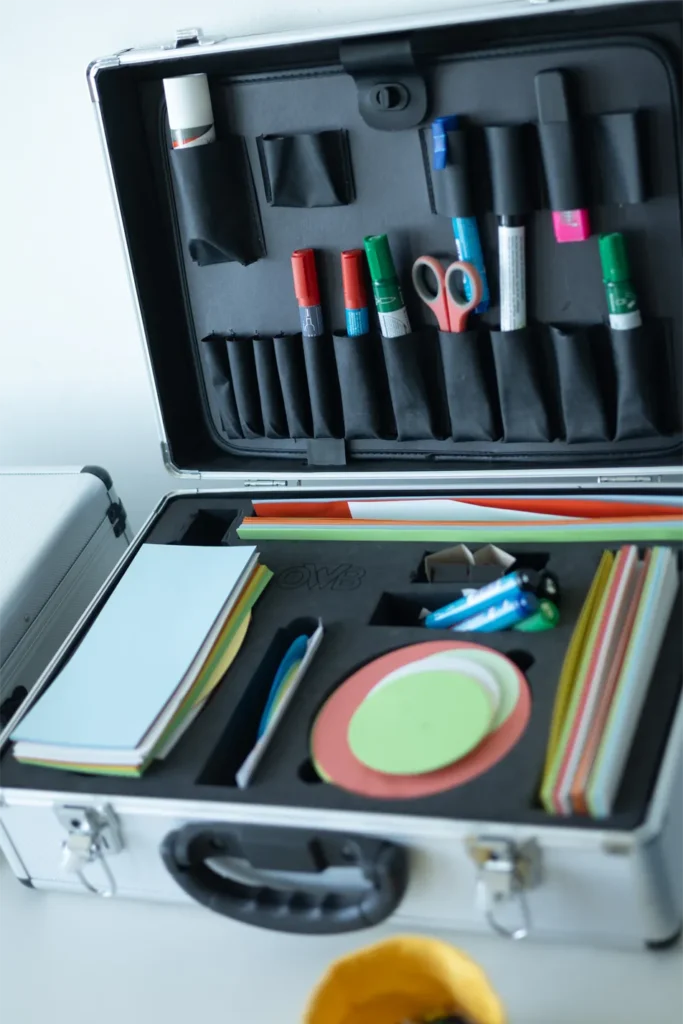 Das Bild zeigt einen geöffneten Koffer voller Präsentationsmaterialien. Auf der Innenseite des Koffers befinden sich verschiedene Fächer, die mit Markern, Scheren und Klebstoff gefüllt sind. Es sind mehrere Buntstifte in unterschiedlichen Farben sowie dicke und dünne Marker zu sehen. Im unteren Bereich liegen verschiedene Papierblätter und bunte Streifen, die für Tagungen verwendet werden können. Zudem sind runde und ovale Papierstücke in verschiedenen Farben vorhanden.