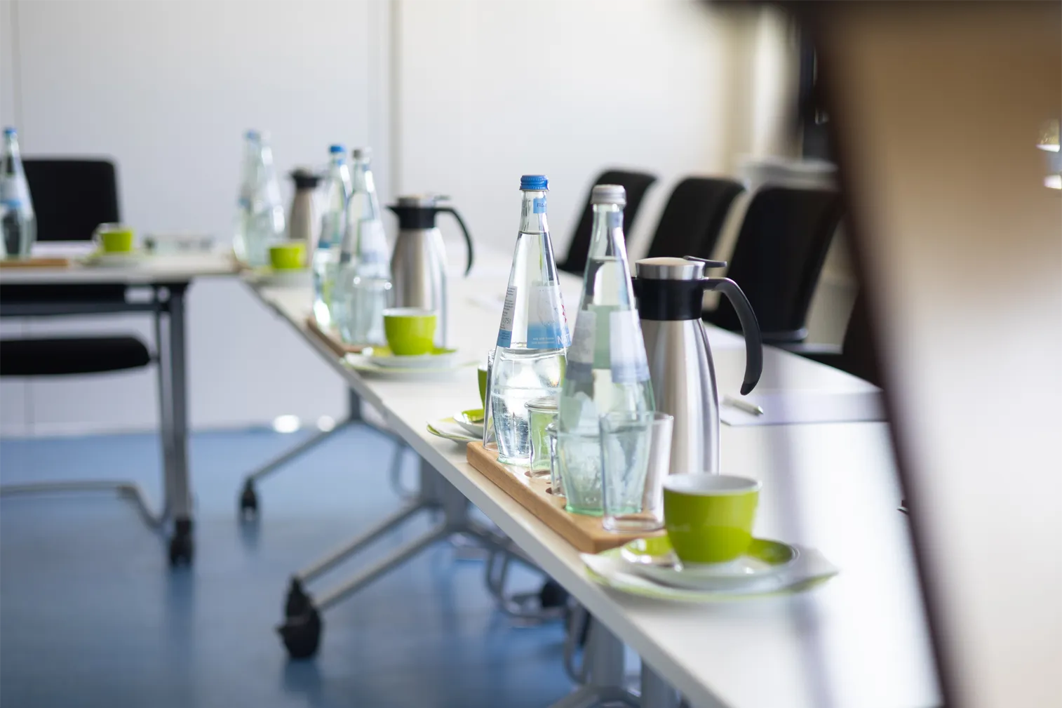 Auf dem Bild ist ein eleganter, aufgeräumter Konferenztisch zu sehen, der mit mehreren Glasflaschen Wasser und Kaffeekannen dekoriert ist. Die Flaschen sind in verschiedenen Größen, einige sind mit klaren Gläsern neben ihnen platziert. Außerdem stehen kleine, grüne Tassen und Untertassen auf dem Tisch, die einen frischen und modernen Eindruck vermitteln. Der Tisch ist in einem hellen Raum angeordnet, während schwarze Stühle um ihn herum stehen