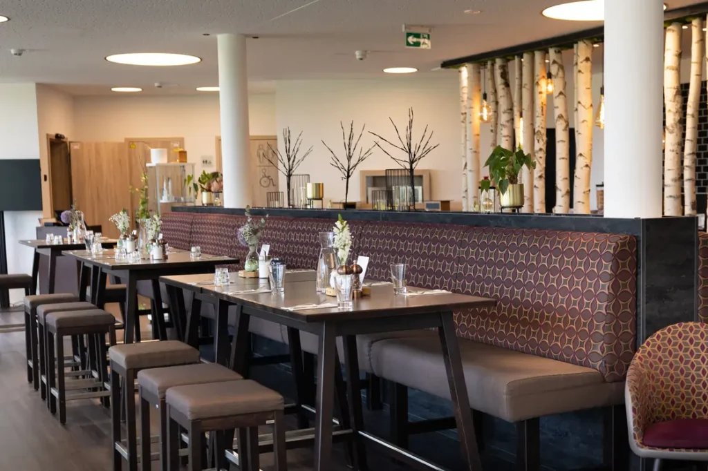 In einem modernen Restaurant sind mehrere Tische mit dunklen Holzoberflächen und gemütlichen Sitzbänken dekoriert. Die Tische sind mit feinen Gläsern, kleinen Blumenarrangements und eleganten Bestecken eingedeckt. Im Hintergrund sind helle Wände und eine auffällige Beleuchtung zu sehen, sowie dekorative Birkenstämme, die eine natürliche Atmosphäre schaffen. Der Raum wirkt freundlich und einladend, ideal für gesellige Treffen oder romantische Abendessen.