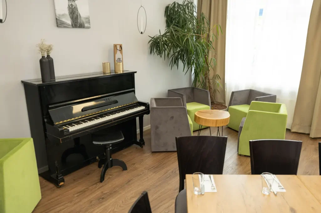 Im Bild ist ein stilvoll eingerichteter Raum zu sehen. In der linken Ecke steht ein schwarzes Klavier mit einer Klavierbank und einer einfachen, eleganten Dekoration auf der Klavieroberfläche. Daneben befinden sich mehrere Sitzgelegenheiten: zwei grau gepolsterte Sessel und zwei leuchtend grüne Sesseln, die um einen kleinen, runden Tisch aus Holz gruppiert sind. Im Hintergrund sieht man eine große Pflanze, die zur angenehmen, einlad