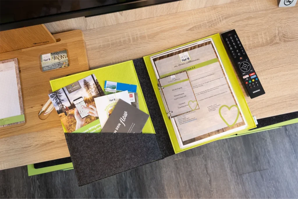 Das Bild zeigt einen Tisch mit verschiedenen Informationsmaterialien für Gäste eines Hotels. Auf einem hellen Holzuntergrund liegt ein Ordner mit einer leuchtend grünen und grauen Hülle, der eine WLAN-Zugangsinformation enthält. Daneben befinden sich mehrere Prospekte und Flyer, unter anderem mit Aktivitäten in der Umgebung und Informationen über das Hotel Fünf 10. Ein Kugelschreiber und eine Notizblock sind ebenfalls sichtbar. Rechts im Bild liegt eine Fernbedienung auf dem