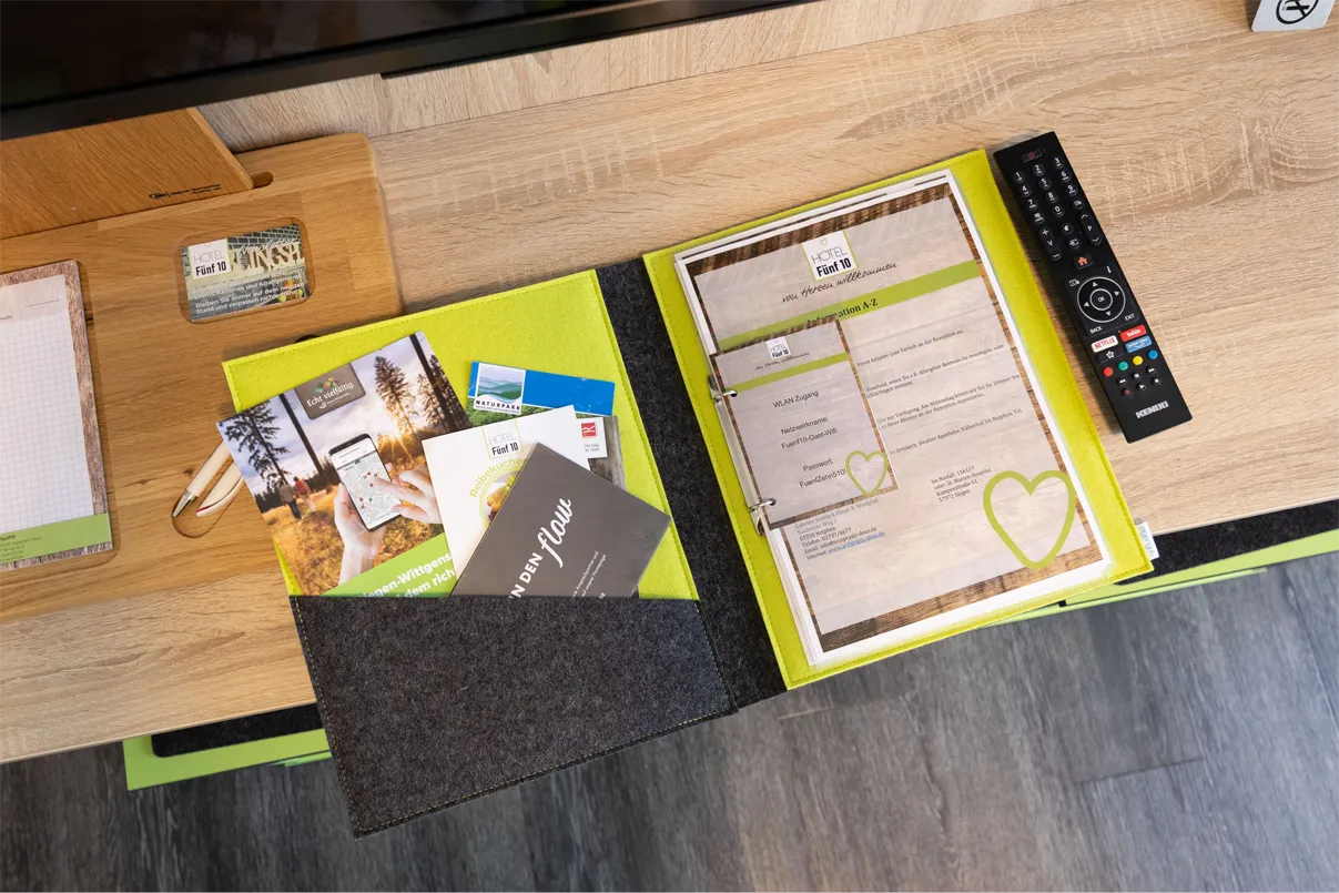 Das Bild zeigt einen Tisch mit verschiedenen Informationsmaterialien für Gäste eines Hotels. Auf einem hellen Holzuntergrund liegt ein Ordner mit einer leuchtend grünen und grauen Hülle, der eine WLAN-Zugangsinformation enthält. Daneben befinden sich mehrere Prospekte und Flyer, unter anderem mit Aktivitäten in der Umgebung und Informationen über das Hotel Fünf 10. Ein Kugelschreiber und eine Notizblock sind ebenfalls sichtbar. Rechts im Bild liegt eine Fernbedienung auf dem