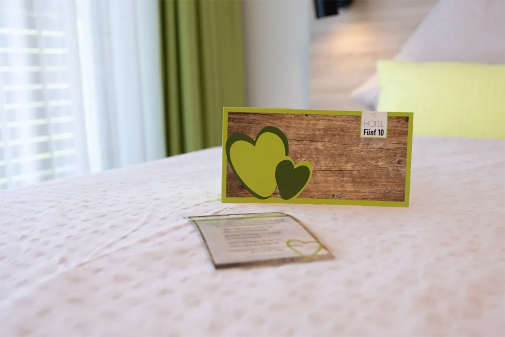 Ein Zimmer im Hotel Fünf 10, auf dem eine grüne Karte mit Herzmotiven und der Aufschrift „Hotel Fünf 10“ liegt. Im Hintergrund sind helle Vorhänge und ein gelbes Kissen sichtbar. Zusätzlich liegt ein Informationsblatt auf dem Bett. Die gesamte Atmosphäre strahlt eine einladende und gemütliche Stimmung aus.