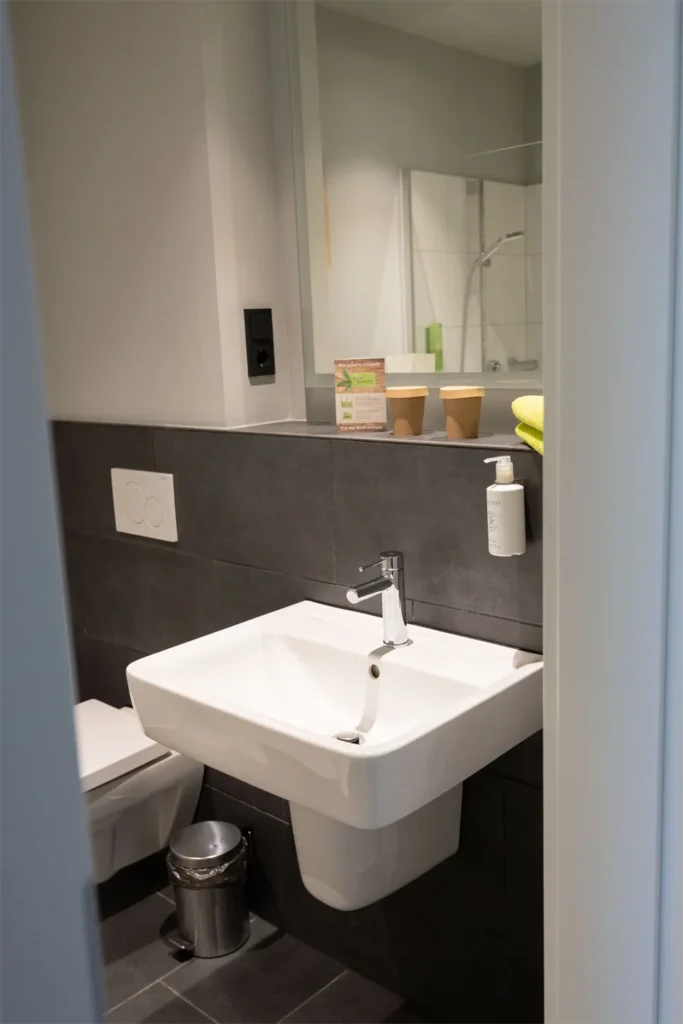 Das Bild zeigt ein modernes Badezimmer mit einem schlichten, weißen Waschbecken, das an der Wand montiert ist. Neben dem Waschbecken befindet sich ein Seifenspender. Im Hintergrund sind zwei Kaffeepötte aus Pappe und ein grünes Handtuch auf einer Ablage sichtbar. An der Wand sind schlichte, dunkle Fliesen verlegt. Zu sehen ist außerdem eine graue Mülltonne, die aus Metall besteht. Der Waschbereich wirkt hell und einlad