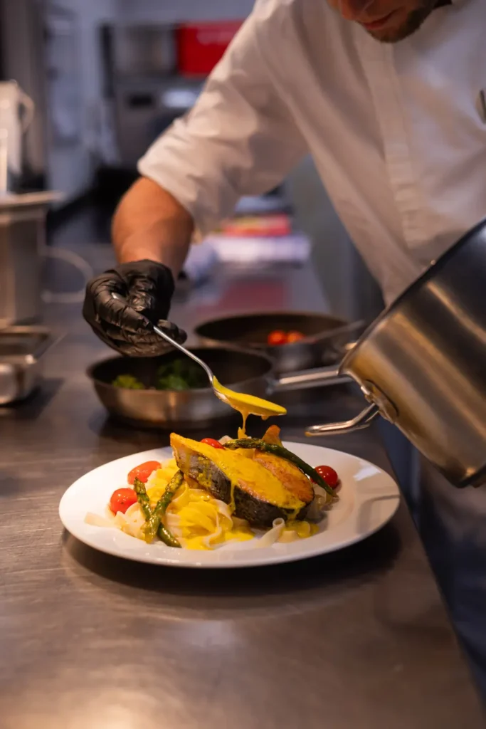Ein Koch in weißer Berufskleidung und schwarzen Handschuhen richtet kunstvoll einen Teller an. Auf dem weißen Teller befindet sich eine Portion Pasta mit einer cremigen, gelben Sauce, neben der farbenfrohe Zutaten wie gegrillte grüner Spargel und Kirschtomaten angerichtet sind. Der Koch gießt gerade eine zusätzliche Sauce über das Gericht, während sich im Hintergrund Küchengeräte und Töpfe befinden, die auf ein aktives Küchenumfeld
