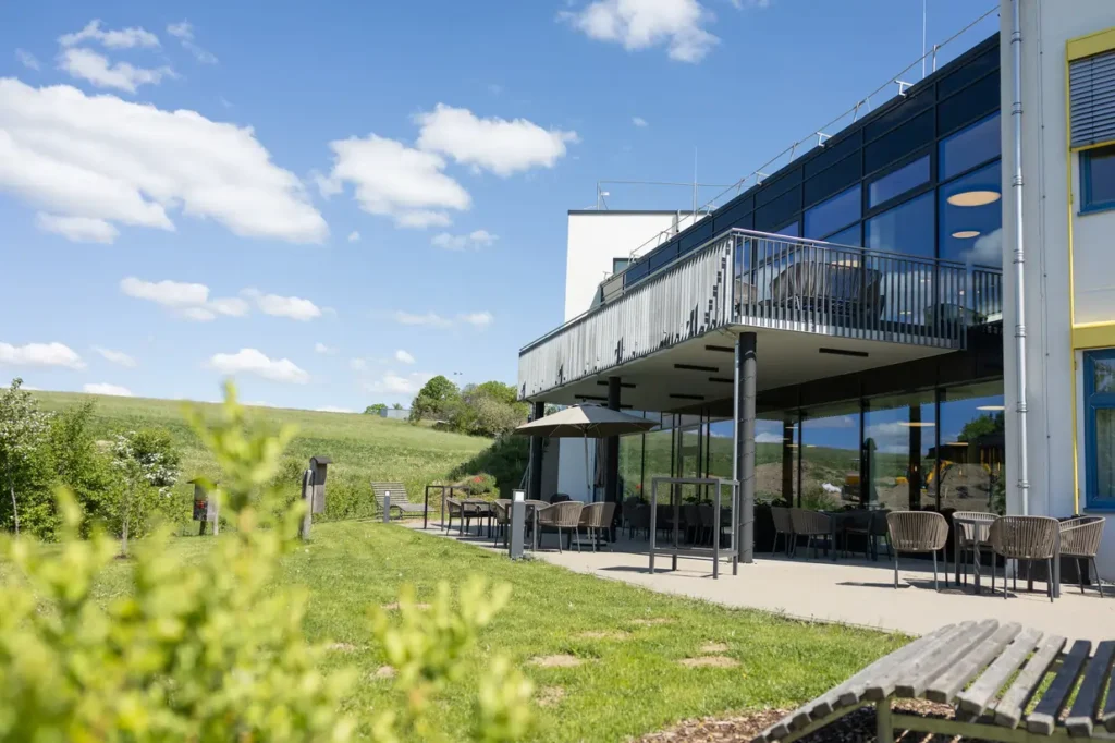 Eine moderne Gebäudeansicht mit großen Glasfenstern und einem Außenbereich. Im Vordergrund befindet sich eine grüne Wiese, die von Sträuchern gesäumt ist. Auf der Terrasse stehen Stühle und Tische sowie ein Sonnenschirm. Der Himmel ist blau mit einigen weißen Wolken, und im Hintergrund sind sanfte Hügel zu sehen.