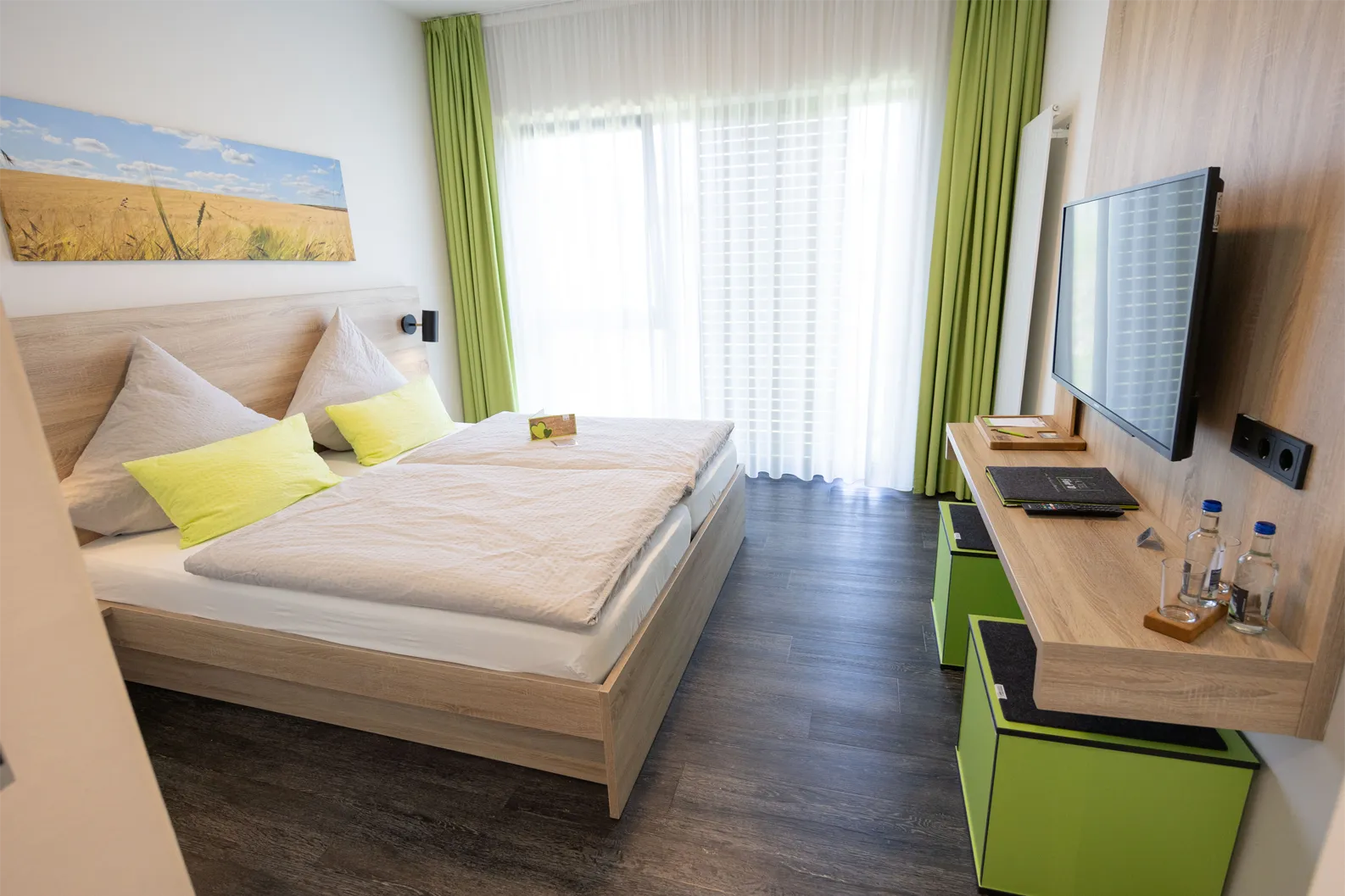 Das Bild zeigt ein modernes Hotelzimmer mit einem großen Doppelbett, das mit einer hellen Bettwäsche und zwei dekorativen Kissen in leuchtendem Gelb ausgestattet ist. An der Wand über dem Bett hängt ein Kunstwerk, das eine Landschaft mit goldenen Feldern und blauem Himmel darstellt. Neben dem Bett stehen zwei kleine Tischlampen. Große Fenster mit hellen, durchsichtigen Vorhängen sorgen für viel Tageslicht. An der Wand gegenüber steht ein Flatscreen-Fernse