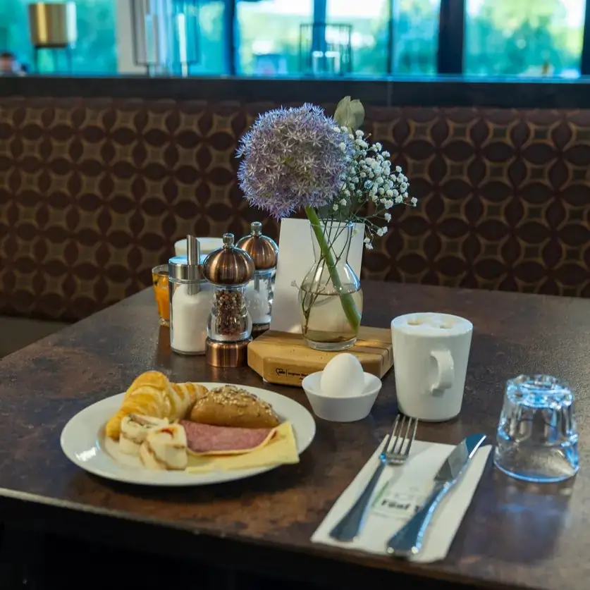 Auf einem rustikalen Tisch im Hotel Fünf10 Restaurant ist ein köstliches Frühstück angerichtet. Auf einem weißen Teller liegen ein Croissant, ein Brötchen, eine Scheibe Wurst und ein Omelett, umrahmt von Scheiben einer Banane. Neben dem Teller befindet sich ein kleines, weißes Schälchen mit einem gekochten Ei. In der Mitte des Tisches steht eine Glasvase mit frischen Blumen, darunter lila Allium und kleine weiße Blüten.