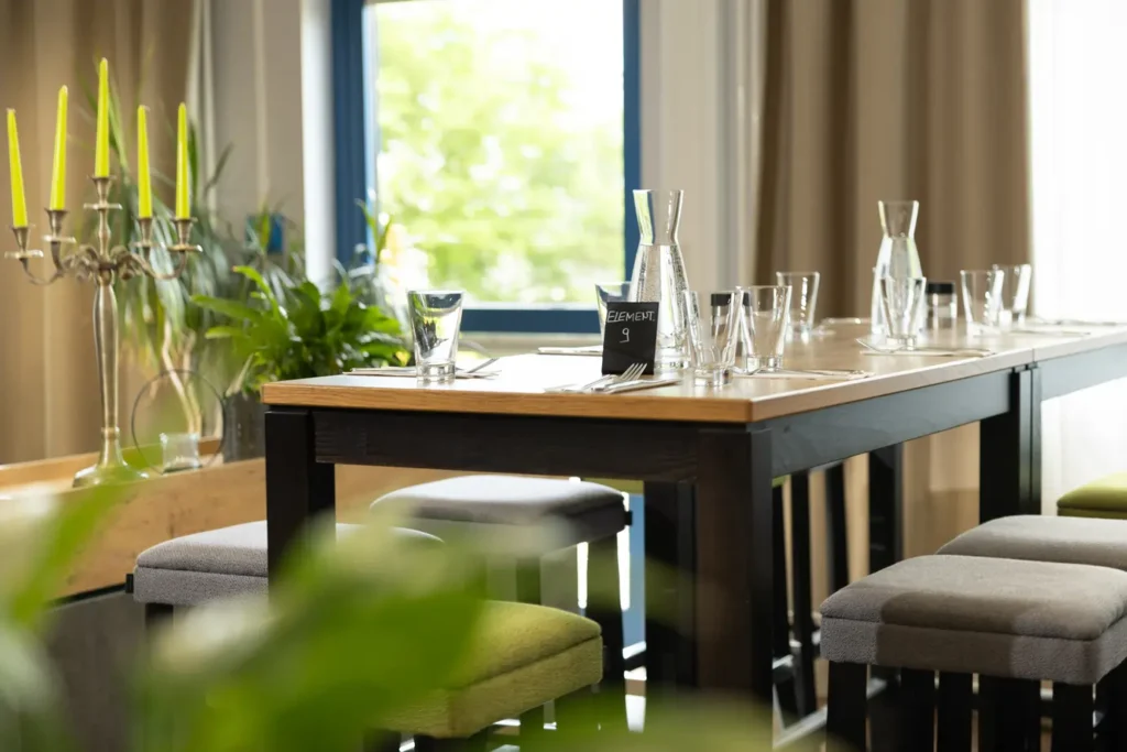 In einem stilvoll eingerichteten Raum steht ein großer Tisch aus Holz mit schwarzen Beinen. Auf dem Tisch sind mehrere Gläser und Krüge Wasser platziert, sowie Besteck und Platzkarten. Im Hintergrund ist ein eleganter Kerzenständer mit leuchtend gelben Kerzen zu sehen, umgeben von grünen Pflanzen, die eine einladende Atmosphäre schaffen. Durch das Fenster fällt Tageslicht und es sind Vorhänge zu erkennen, die den Raum gemütlich wirken lassen.