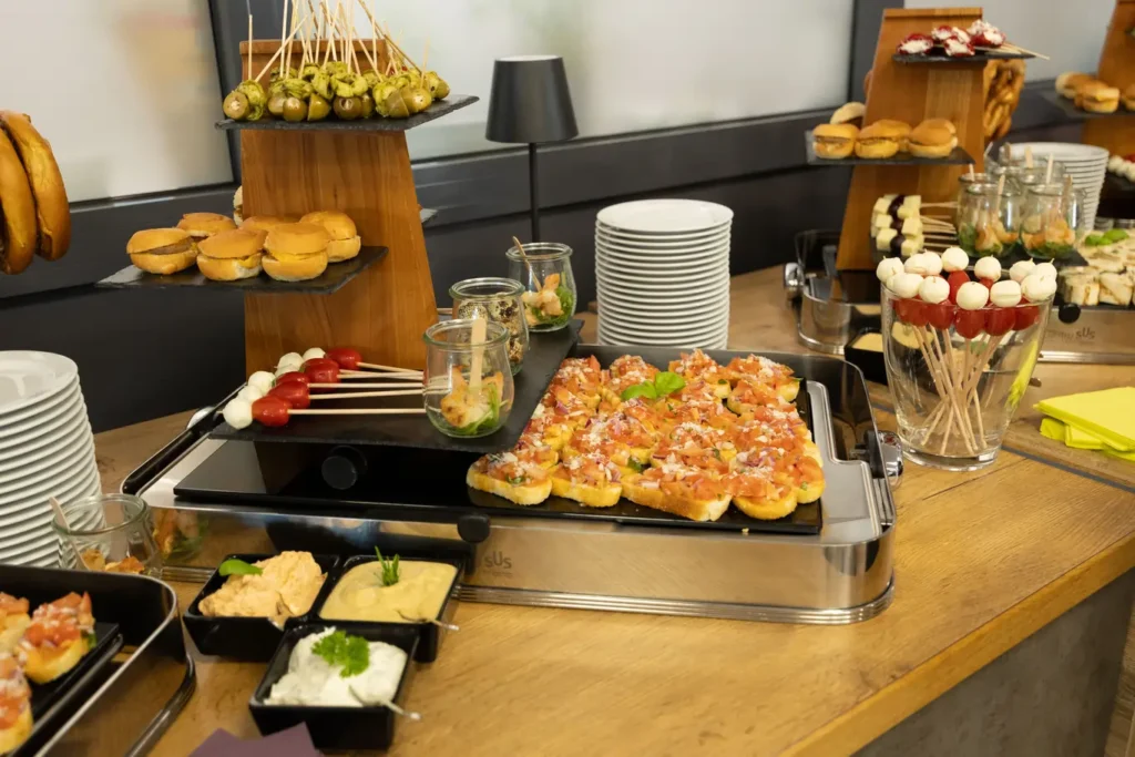 Ein vielfältiges Buffet mit kleinen Speisen: Auf einer Holzplatte befinden sich Brötchen und verschiedene Spieße mit Oliven und Tomaten. In Gläsern sind frische Salate und Dips platziert, während auf einer anderen Platte belegte Brote mit Tomaten und Käse angeboten werden. Im Hintergrund sind Stapel von Tellern sowie weitere Fingerfood-Optionen wie Käsewürfel und süße Leckereien zu sehen. Die Tischoberfläche ist aus Holz