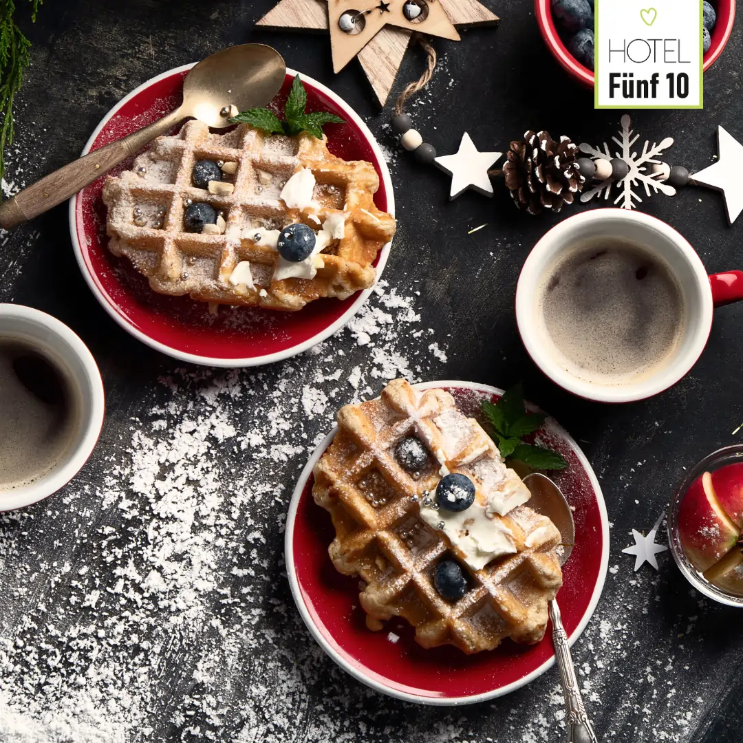 Zwei Teller mit frisch gebackenen Waffeln, garniert mit Blaubeeren und Sahne, serviert mit Tassen Kaffee auf weihnachtlich dekoriertem Tisch im Hotel Fünf10
