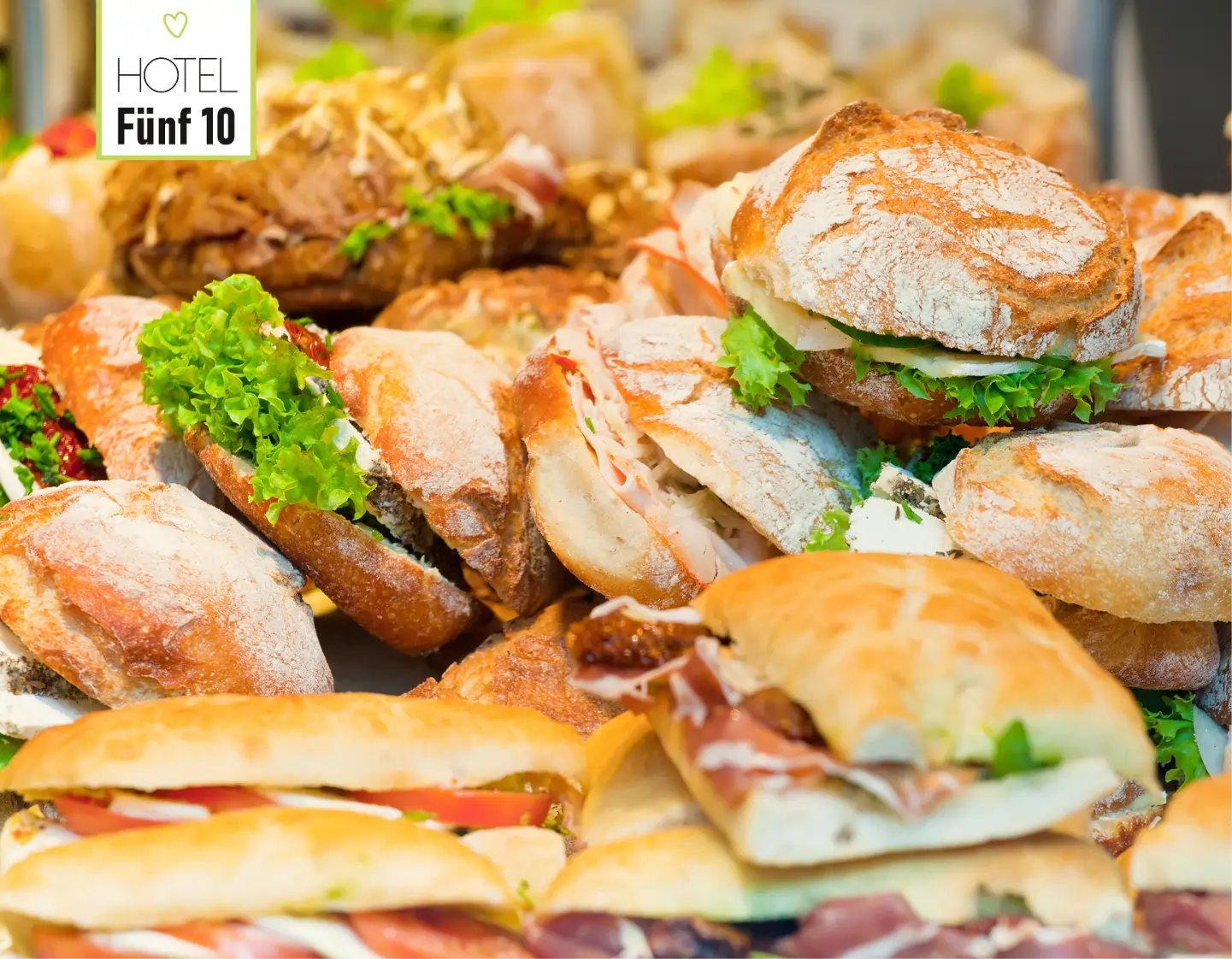 Verschiedene frische belegte Brötchen mit Salat, Käse und Aufschnitt auf einem Buffet, im Hintergrund das Logo des Hotels Fünf 10